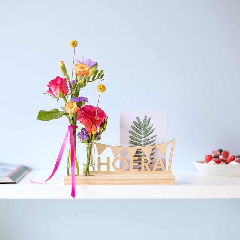 Brievenbuspakket met een houten plankje, houten hoera tekst en twee vaasjes met verse bloemen. Er is ruimte voor een wenskaart in de plank