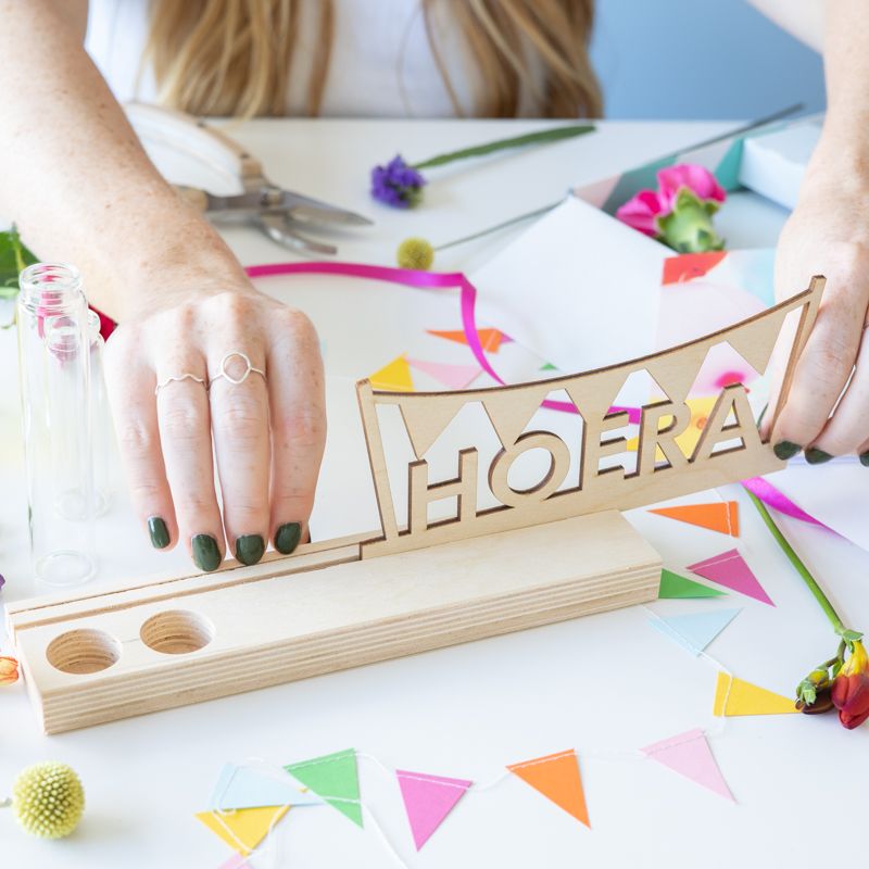Brievenbuspakket met een houten plankje, houten hoera tekst en twee vaasjes met verse bloemen. Er is ruimte voor een wenskaart in de plank