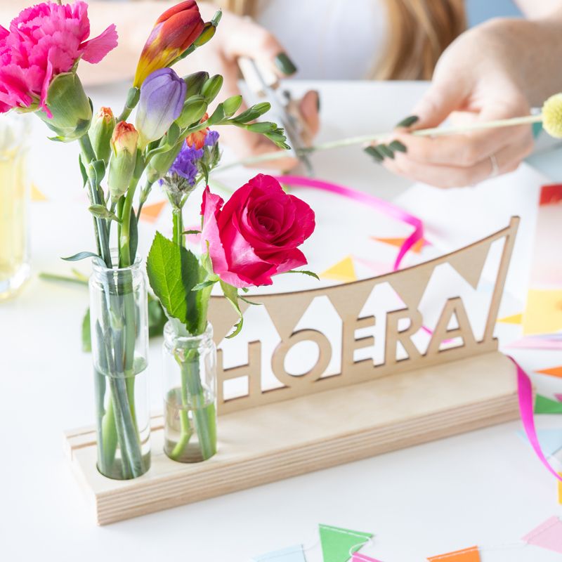 Brievenbuspakket met een houten plankje, houten hoera tekst en twee vaasjes met verse bloemen. Er is ruimte voor een wenskaart in de plank