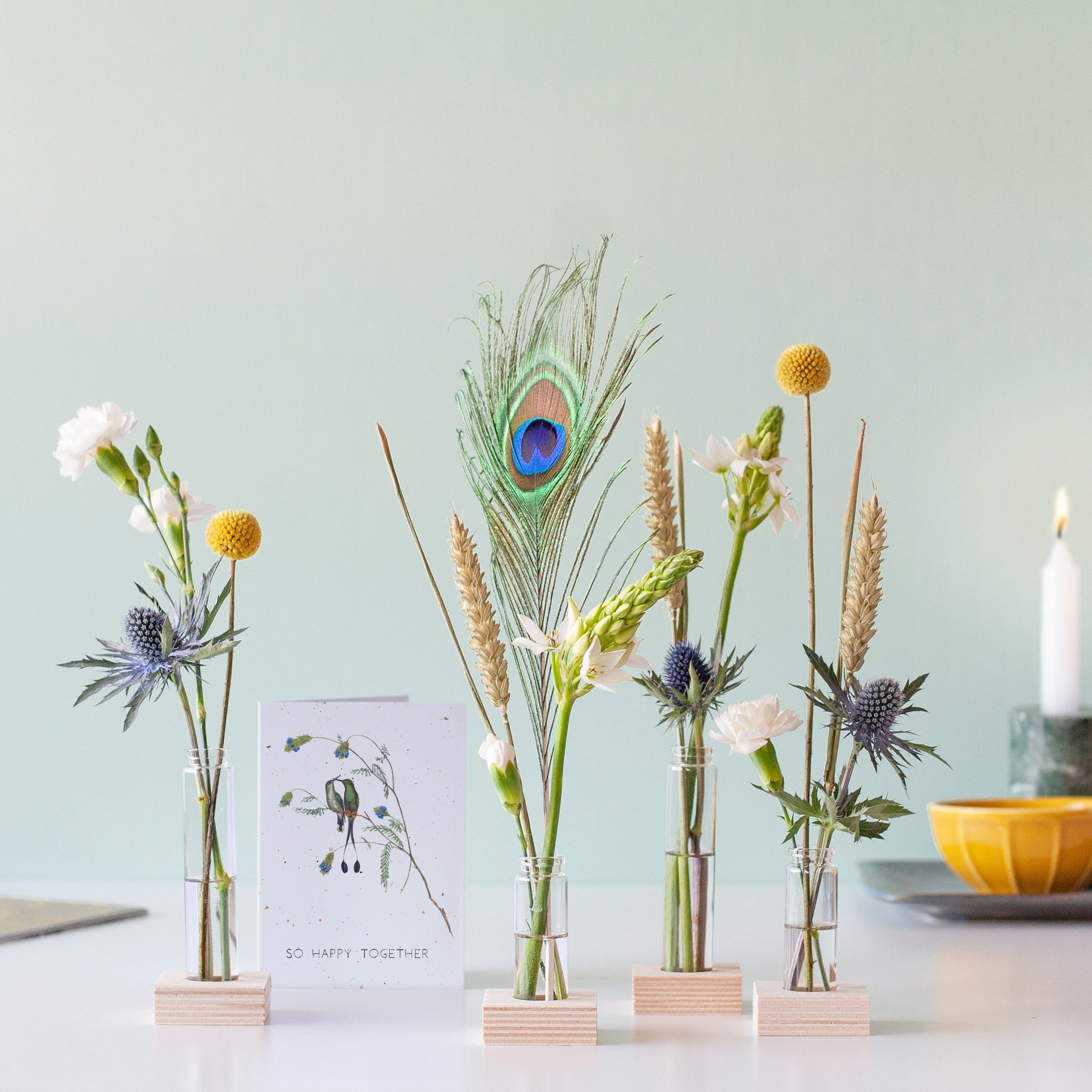 Brievenbuspakketje met vier vaasjes in houten blokjes met verschillende bloemen en een pauwenveer