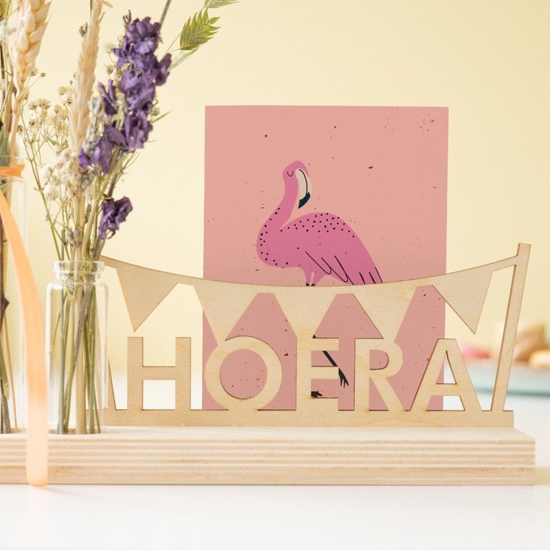 houten plankje met daarop een houten hoera tekst en vlaggenlijn en daarnaast een vaasje met droogbloemen in natuurtinten met paarse bloemetjes