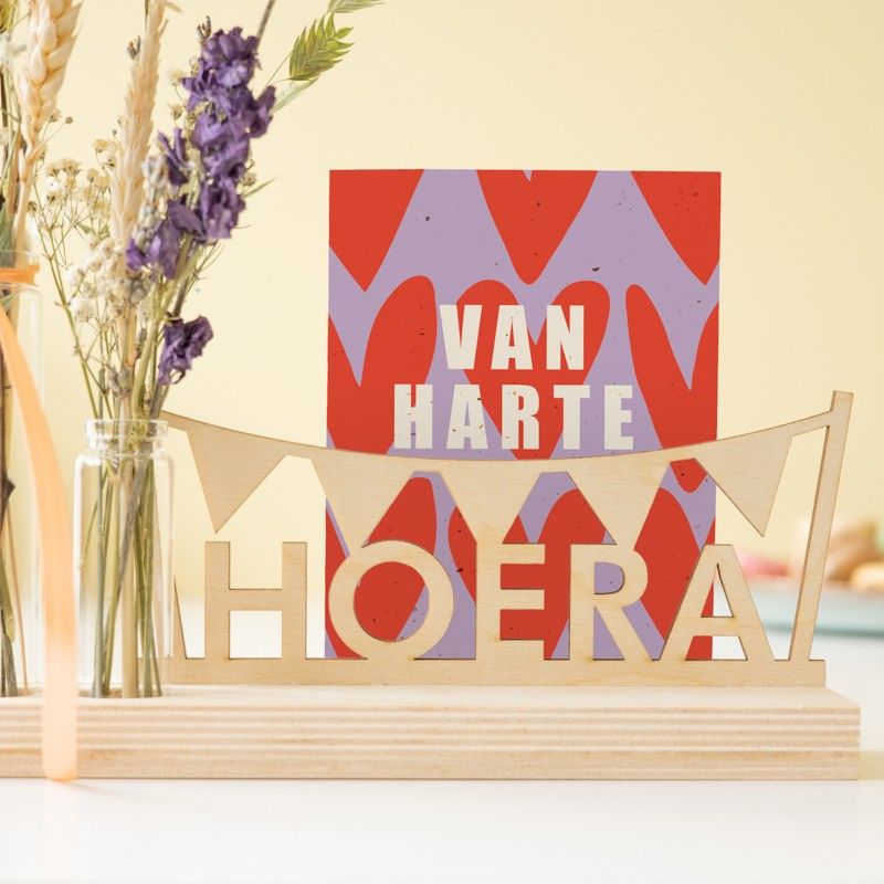 houten plankje met daarop een houten hoera tekst en vlaggenlijn en daarnaast een vaasje met droogbloemen in natuurtinten met paarse bloemetjes