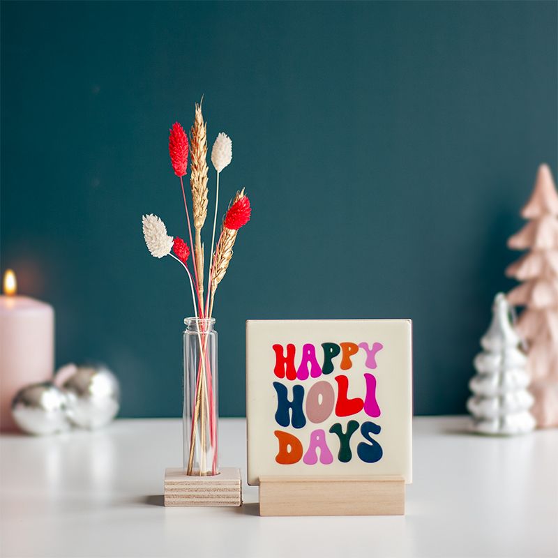 Cremekleurig tegeltje met in verschillende kleuren Happy holidays geschreven. Het tegeltje staat op een houten houdertje. Daarnaast staat een vaasje met droogbloemen in rood en creme kleuren. Het vaasje staat in een vierkant houten houdertje