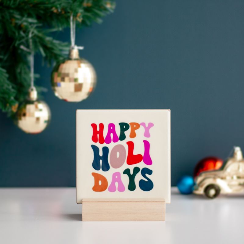 Cremekleurig tegeltje met in verschillende kleuren de tekst happy holidays. De tegel staat in een houten houdertje.