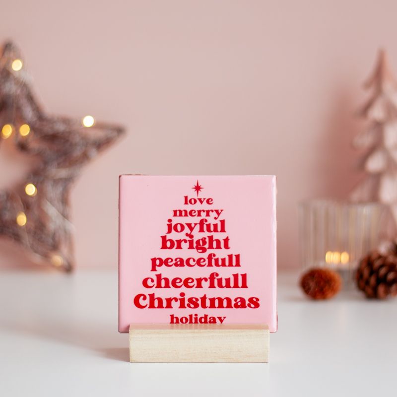 Licht roze tegeltje met in het rood de tekst love merry joyful bright peacefull cheerfull christmas holiday. De woorden staan onder elkaar en vormen een kerstboom. Boven het eerste woord staat een ster. Het tegeltje staat in een houten houder. 