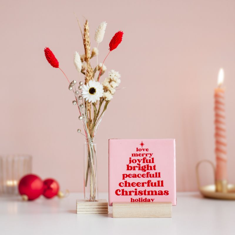 Licht roze tegeltje met in het rood de tekst love merry joyful bright peacefull cheerfull christmas holiday. De woorden staan onder elkaar en vormen een kerstboom. Boven het eerste woord staat een ster. Het tegeltje staat in een houten houder. Daarnaast staat een vaasje in een houten houdertje met daarin droogbloemen in rood en witte kleuren.