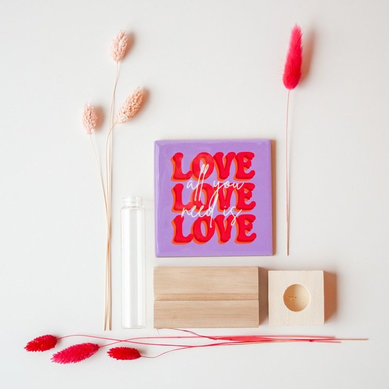 Paars tegeltje met All you need is love love love met houten standaard liggend op een witte ondergrond. Ernaast rode en naturel droogbloemen, glazen vaasje en houten houder voor het vaasje.