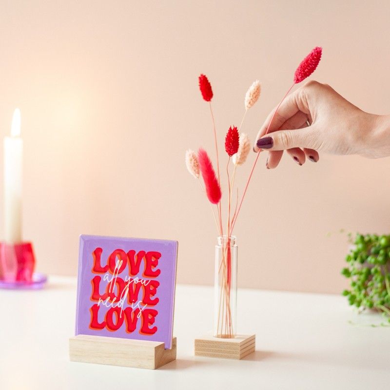 Paars tegeltje met de tekst "All you need is love love love", geplaatst in een houten standaard. Een hand steekt een droogbloem in een klein glazen vaasje naast het tegeltje.