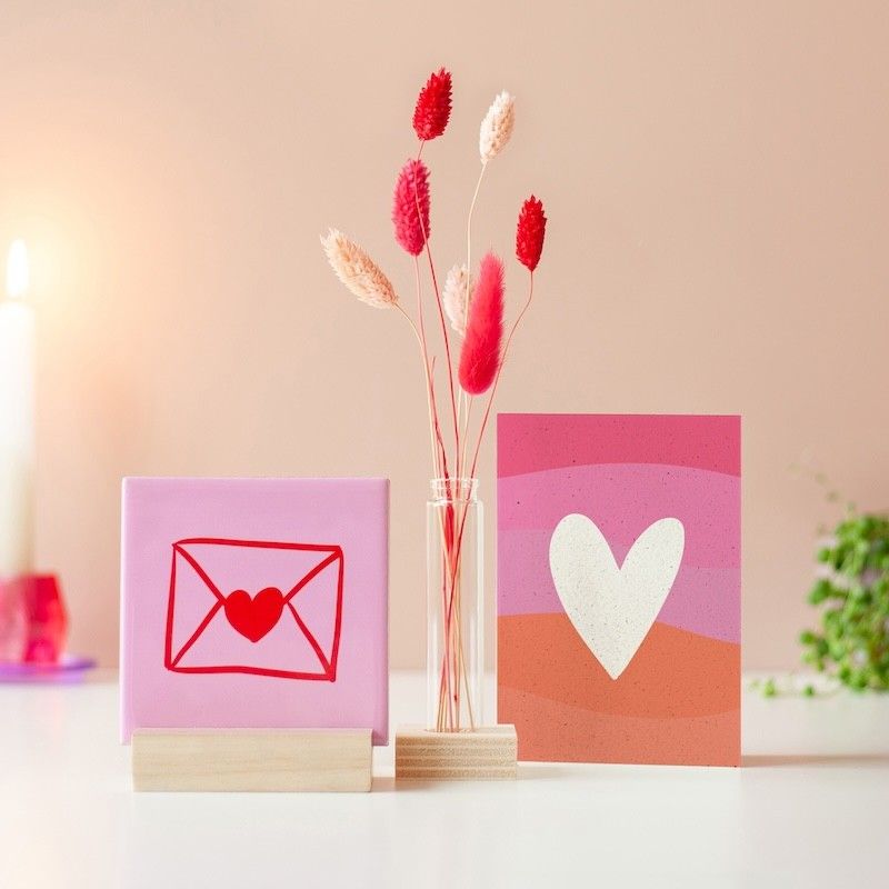 Roze tegeltje met een rode illustratie van een envelop met hart, in houten standaard. Ernaast een glazen vaasje in een houten standaard met roze en beige droogbloemen. Daarnaast staat een roze oranje wenskaart met een wit hart erop.