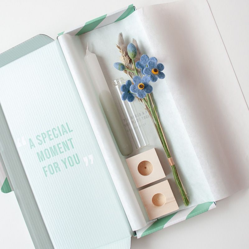 Een groen witte kaars met daarnaast een vaasje met blauwe vilten bloemetjes