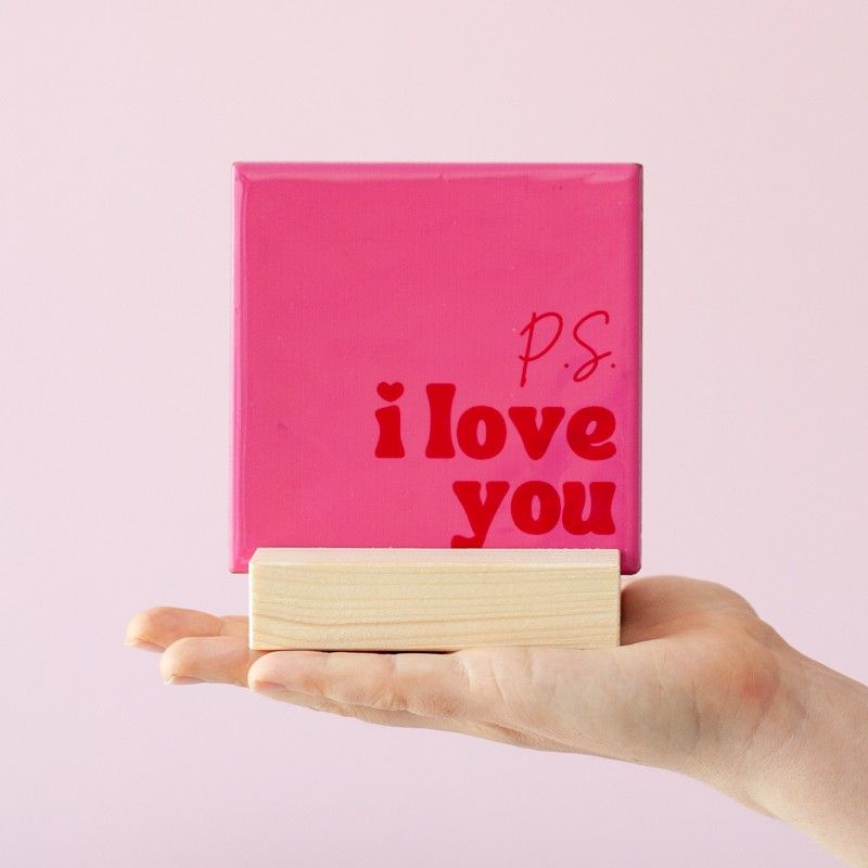 Een hand waar een roze tegeltje met P.S. I love you in rode letters in een houten standaard op staat