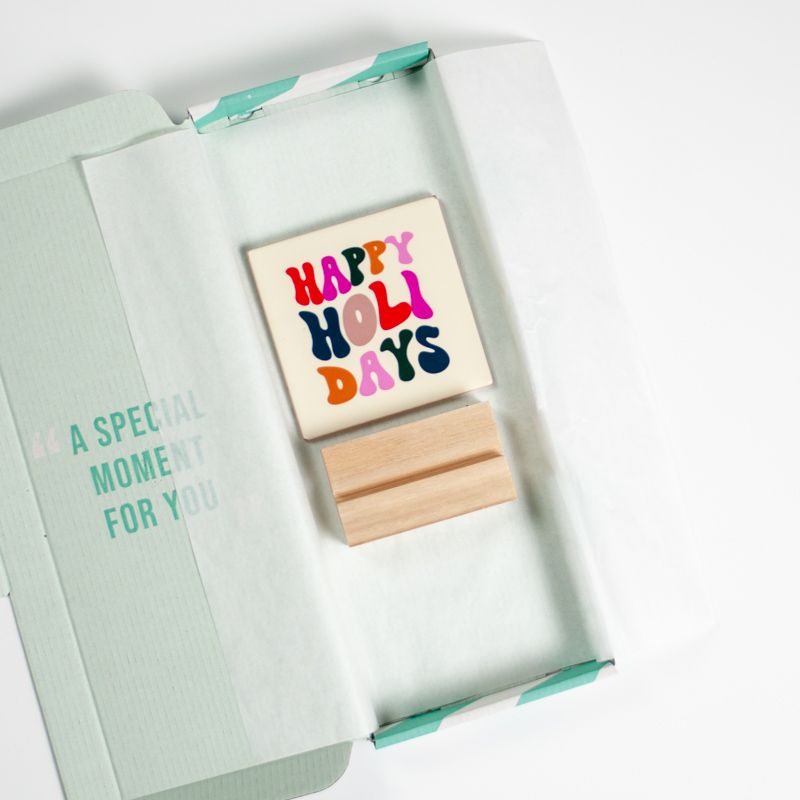 Cremekleurig tegeltje met in verschillende kleuren de tekst happy holidays. De tegel staat in een houten houdertje.