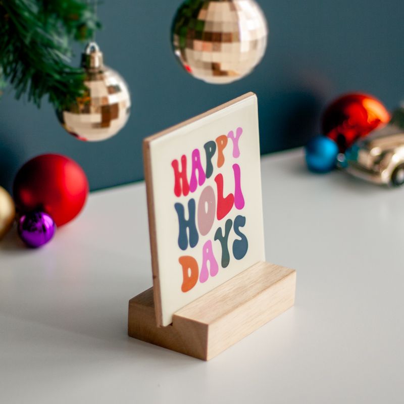 Cremekleurig tegeltje met in verschillende kleuren de tekst happy holidays. De tegel staat in een houten houdertje.