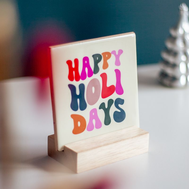 Cremekleurig tegeltje met in verschillende kleuren Happy holidays geschreven. Het tegeltje staat op een houten houdertje. Daarnaast staat een vaasje met droogbloemen in rood en creme kleuren. Het vaasje staat in een vierkant houten houdertje