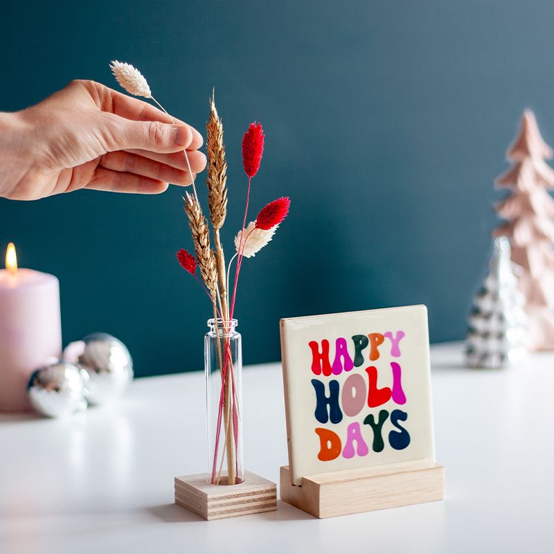 Cremekleurig tegeltje met in verschillende kleuren Happy holidays geschreven. Het tegeltje staat op een houten houdertje. Daarnaast staat een vaasje met droogbloemen in rood en creme kleuren. Het vaasje staat in een vierkant houten houdertje