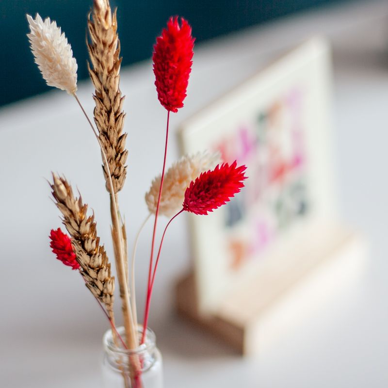Cremekleurig tegeltje met in verschillende kleuren Happy holidays geschreven. Het tegeltje staat op een houten houdertje. Daarnaast staat een vaasje met droogbloemen in rood en creme kleuren. Het vaasje staat in een vierkant houten houdertje