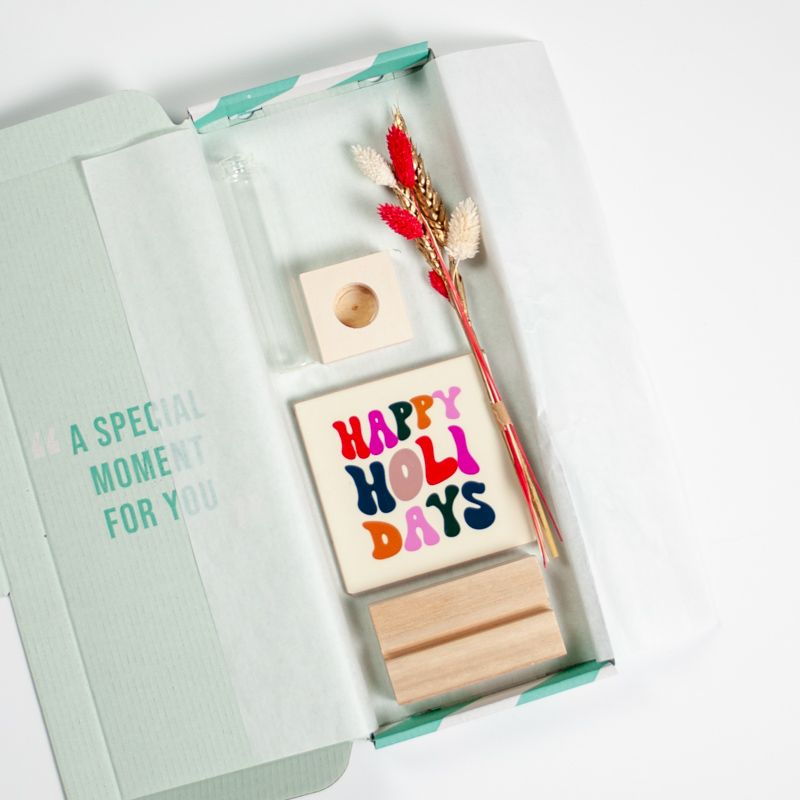 Cremekleurig tegeltje met in verschillende kleuren Happy holidays geschreven. Het tegeltje staat op een houten houdertje. Daarnaast staat een vaasje met droogbloemen in rood en creme kleuren. Het vaasje staat in een vierkant houten houdertje