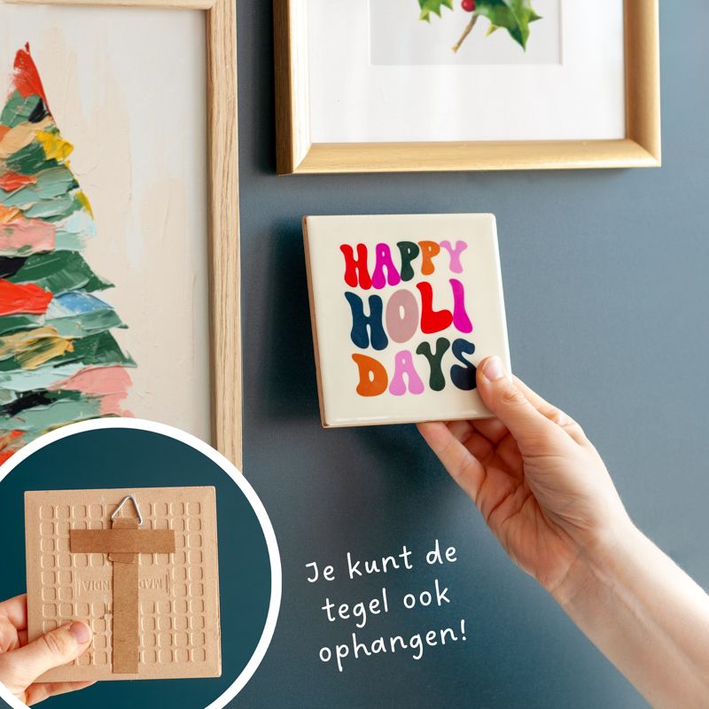 Cremekleurig tegeltje met in verschillende kleuren Happy holidays geschreven. Het tegeltje staat op een houten houdertje. Daarnaast staat een vaasje met droogbloemen in rood en creme kleuren. Het vaasje staat in een vierkant houten houdertje