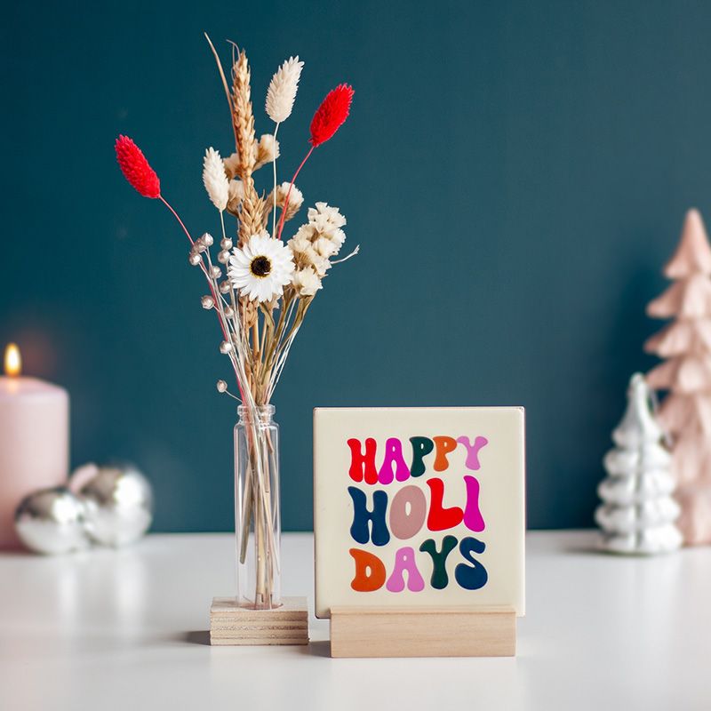 Cremekleurig tegeltje met in verschillende kleuren Happy holidays geschreven. Het tegeltje staat op een houten houdertje. Daarnaast staat een vaasje met droogbloemen in rood en creme kleuren. Het vaasje staat in een vierkant houten houdertje