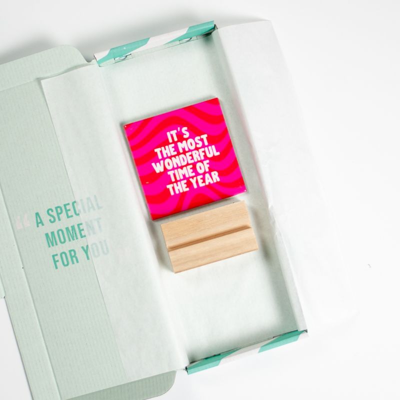 Kersttegel met de tekst "It's the most wonderful time of the year" in witte letters op een roze met rode achtergrond met golvend patroon. De tegel staat op een houten standaard.