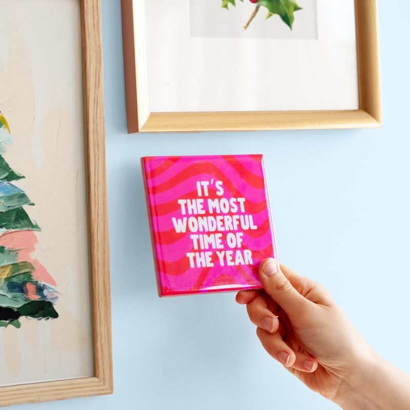 Kersttegel met de tekst "It's the most wonderful time of the year" in witte letters op een roze met rode achtergrond met golvend patroon. De tegel staat op een houten standaard.