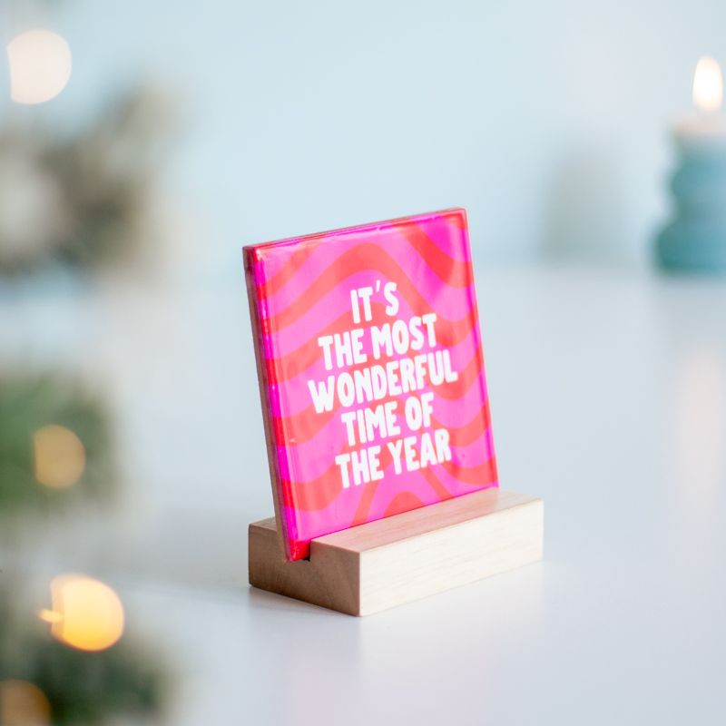 Kersttegel met de tekst "It's the most wonderful time of the year" in witte letters op een roze met rode achtergrond met golvend patroon. De tegel staat op een houten standaard.