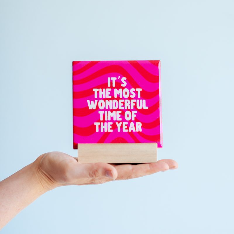 Kersttegel met de tekst "It's the most wonderful time of the year" in witte letters op een roze met rode achtergrond met golvend patroon. De tegel staat op een houten standaard.