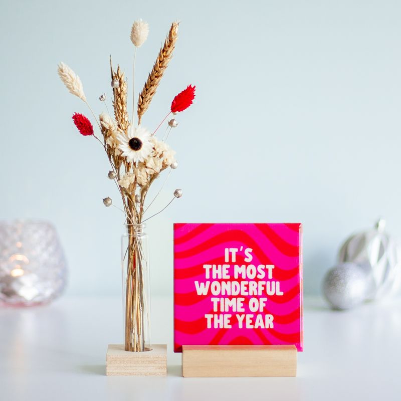 Kersttegel met de tekst "It's the most wonderful time of the year" in witte letters op een roze-rode golvende achtergrond. De tegel staat op een houten standaard naast een vaasje met droogbloemen in rood en wit kleuren.
