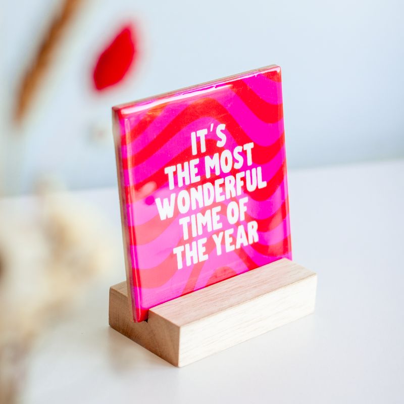 Kersttegel met de tekst "It's the most wonderful time of the year" in witte letters op een roze-rode golvende achtergrond. De tegel staat op een houten standaard naast een vaasje met droogbloemen in rood en wit kleuren.