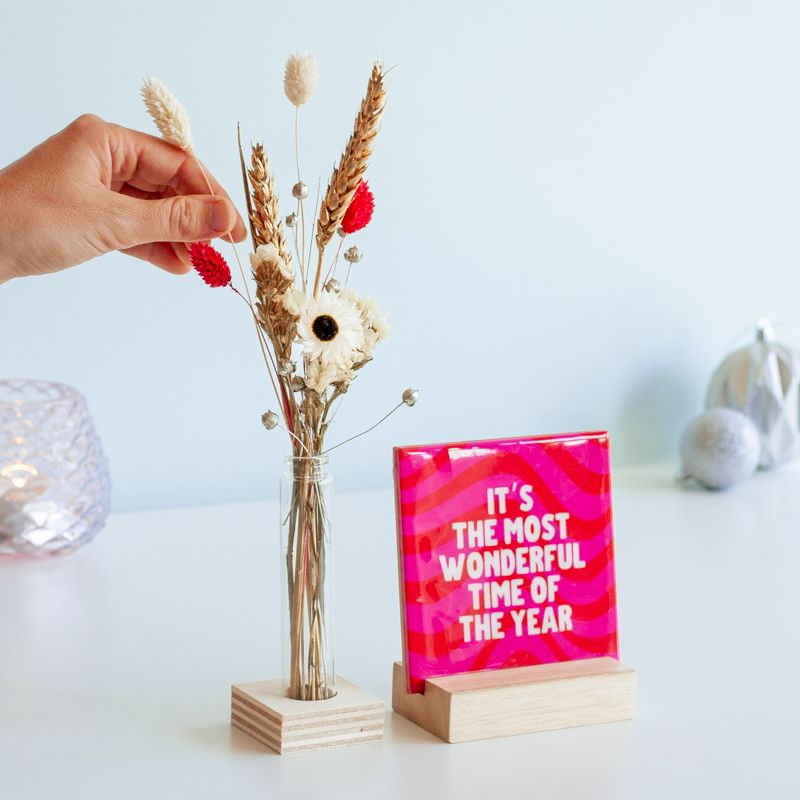 Kersttegel met de tekst "It's the most wonderful time of the year" in witte letters op een roze-rode golvende achtergrond. De tegel staat op een houten standaard naast een vaasje met droogbloemen in rood en wit kleuren.