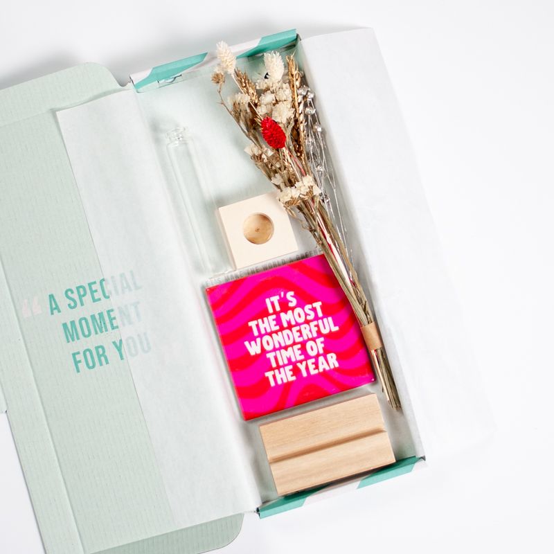 Kersttegel met de tekst "It's the most wonderful time of the year" in witte letters op een roze-rode golvende achtergrond. De tegel staat op een houten standaard naast een vaasje met droogbloemen in rood en wit kleuren.