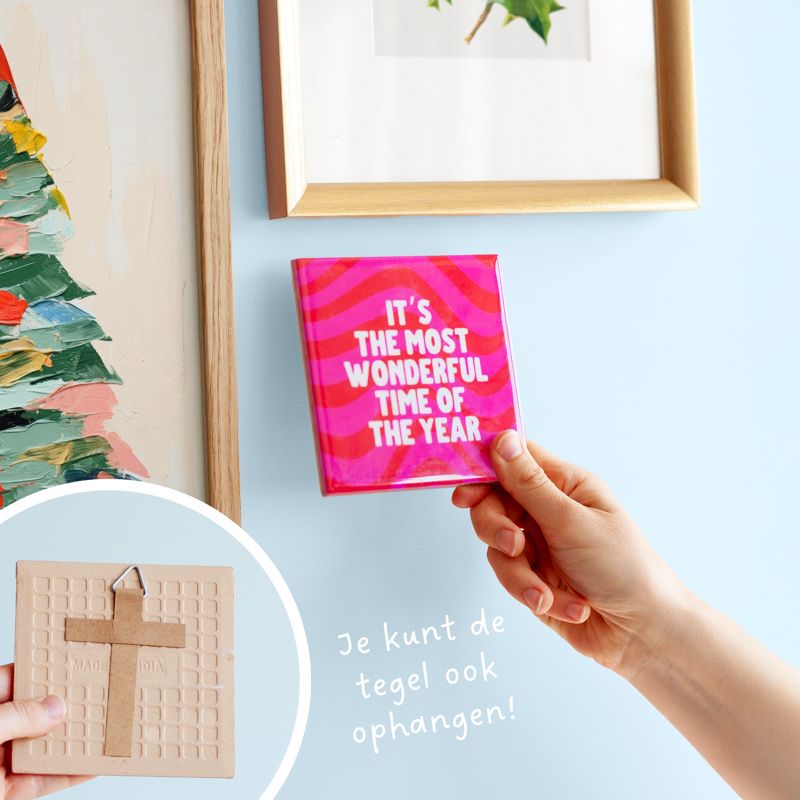 Kersttegel met de tekst "It's the most wonderful time of the year" in witte letters op een roze-rode golvende achtergrond. De tegel staat op een houten standaard naast een vaasje met droogbloemen in rood en wit kleuren.