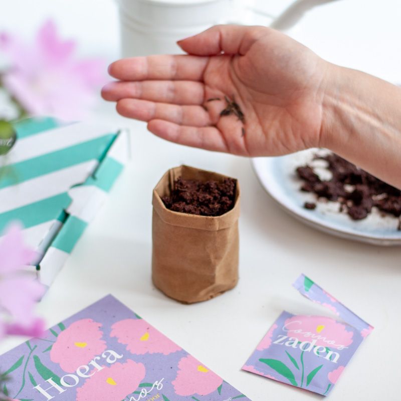  Een hand strooit alle bloemenzaadjes in een mini leatherbag gevuld met aarde. 
