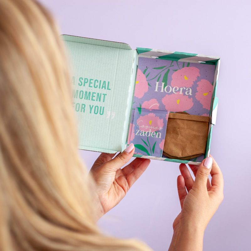 Vrouw houdt het brievenbuscadeautje vast. Je ziet de inhoud: een zakje Hoera zaden, een samengeperste grondtablet en een mini leren potje.