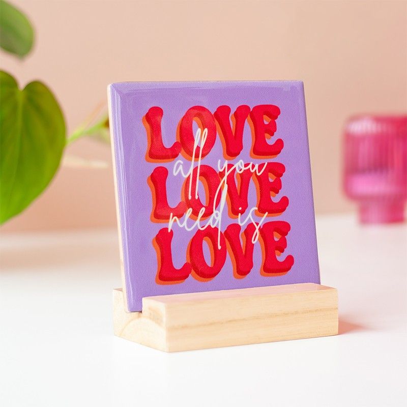 Tegeltje met All you need is love erop. Het tegeltje is paars met roze letters en staat in een houten houdertje