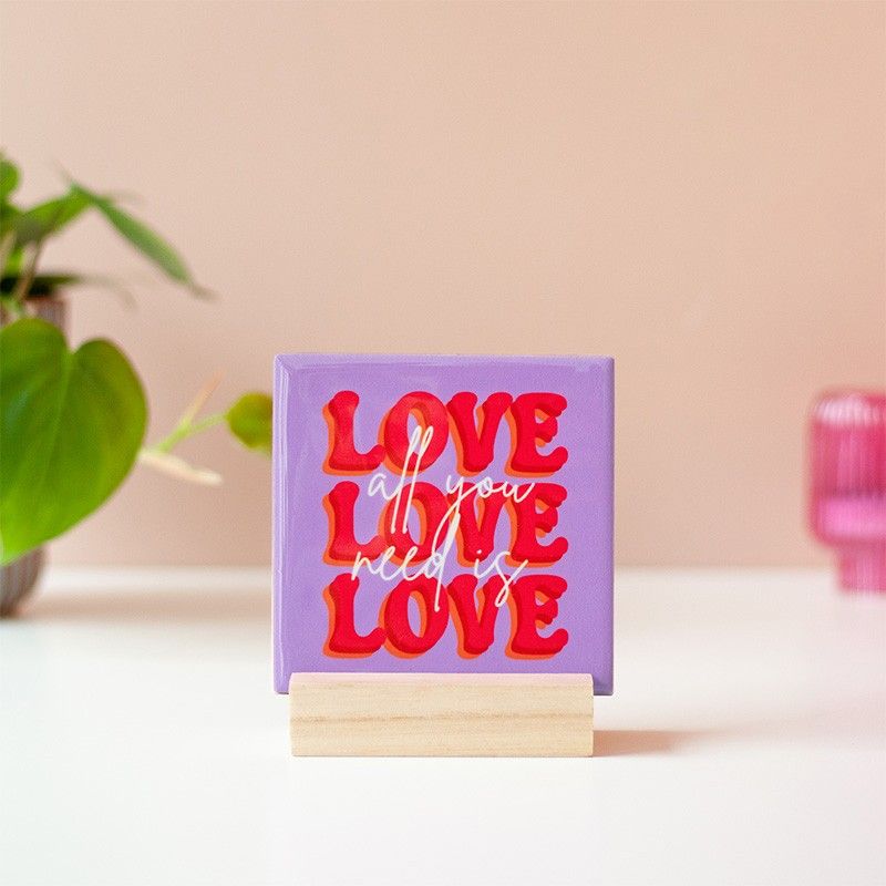 Een vrouw hangt het paarse tegeltje met rode tekst 'LOVE' en witte siertekst 'all you need is love aan de muur.