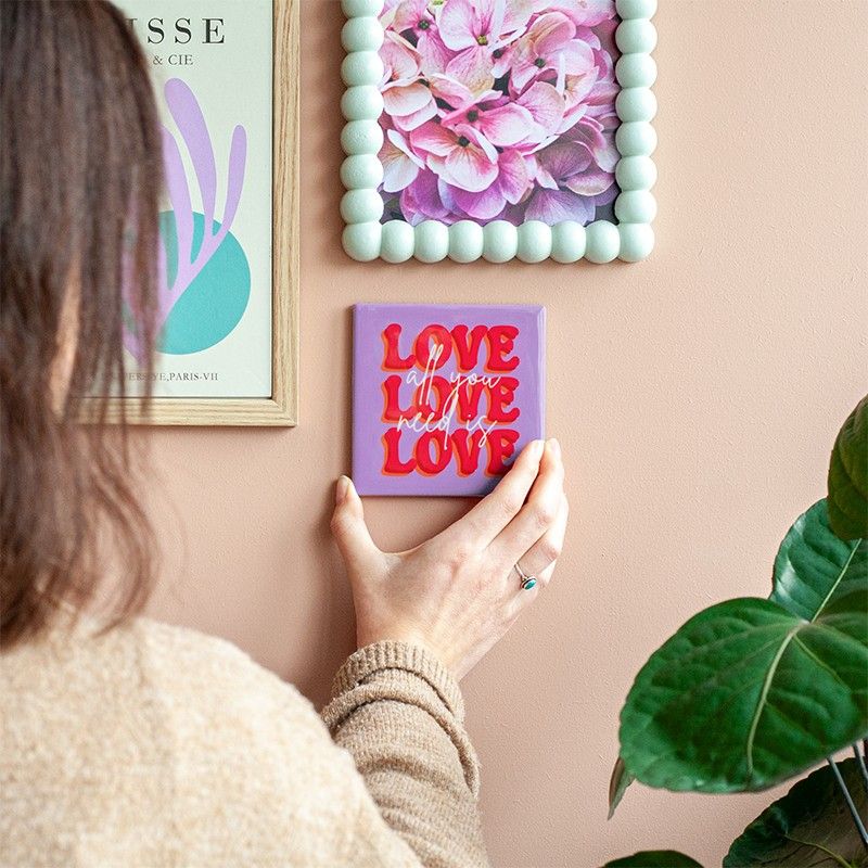 Een vrouw hangt het paarse tegeltje met rode tekst 'LOVE' en witte siertekst 'all you need is love aan de muur.