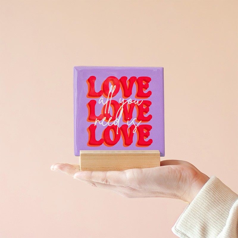 Een hand houdt het tegeltje met "All you need is love love love" in een houten houder vast.