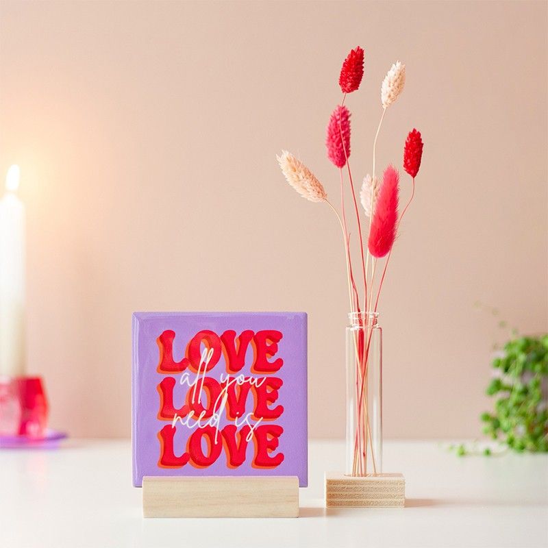 Paars tegeltje met All you need is love love love in een houten standaard met rood en naturel droogbloemen ernaast.