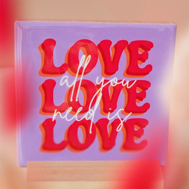 Paars tegeltje met rode tekst 'LOVE' en witte siertekst 'all you need is', in houten standaard.