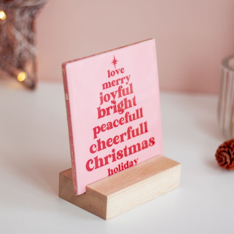Licht roze tegeltje met in het rood de tekst love merry joyful bright peacefull cheerfull christmas holiday. De woorden staan onder elkaar en vormen een kerstboom. Boven het eerste woord staat een ster. Het tegeltje staat in een houten houder. 