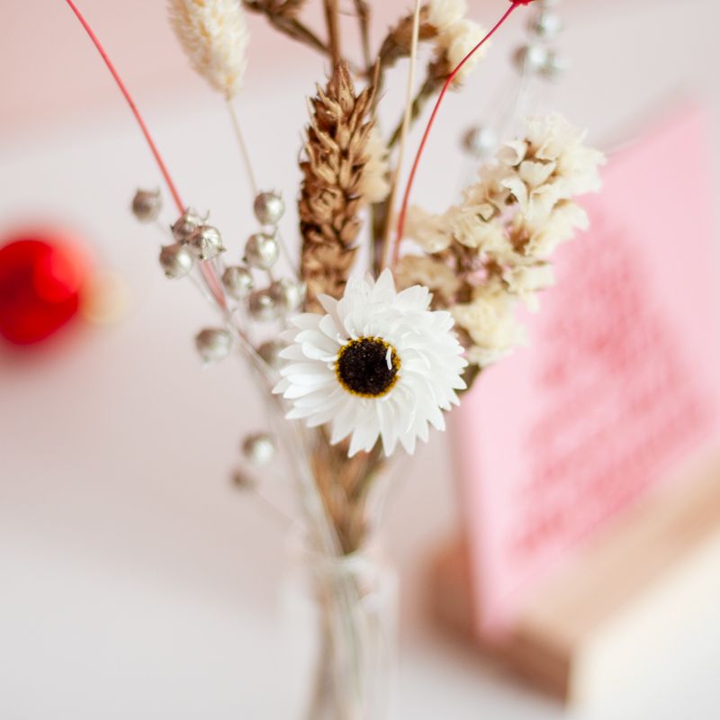 Roze kersttegel met in het rood gelukswensen in kerstboomvorm geschreven met een vaasje droogbloemen ernaast. Beiden staan in een houten blokje