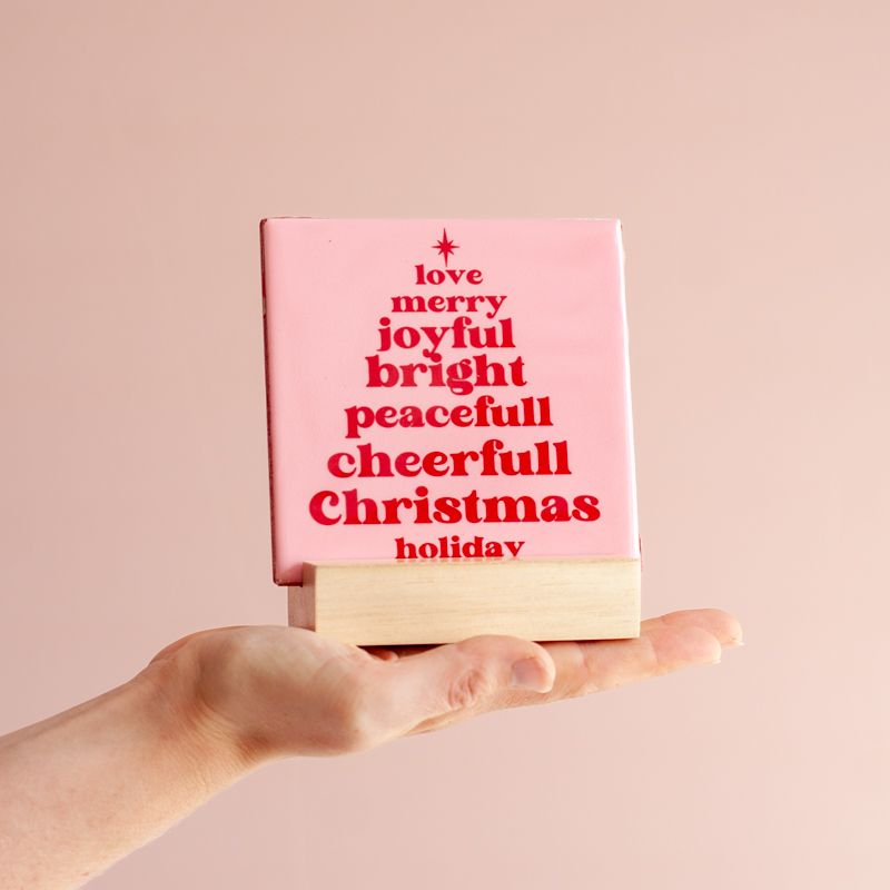 Licht roze tegeltje met in het rood de tekst love merry joyful bright peacefull cheerfull christmas holiday. De woorden staan onder elkaar en vormen een kerstboom. Boven het eerste woord staat een ster. Het tegeltje staat in een houten houder. 