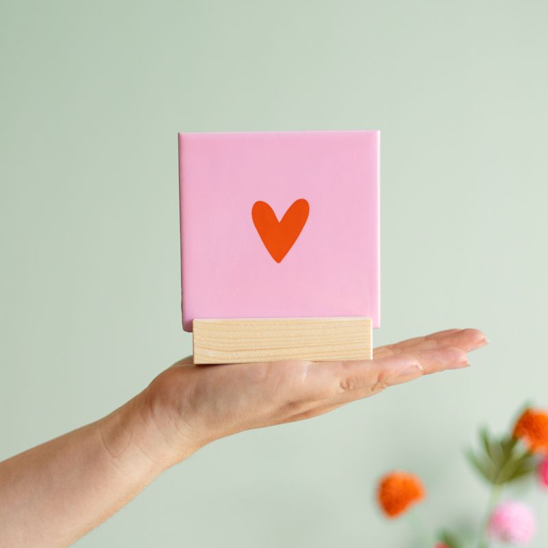 Roze tegeltje met in het midden een klein oranje hartje en het tegeltje staat op een houten houdertje