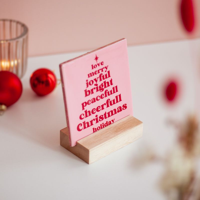 Roze kersttegel met in het rood gelukswensen in kerstboomvorm geschreven met een vaasje droogbloemen ernaast. Beiden staan in een houten blokje