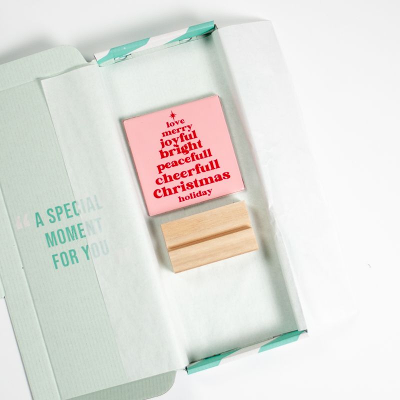 Licht roze tegeltje met in het rood de tekst love merry joyful bright peacefull cheerfull christmas holiday. De woorden staan onder elkaar en vormen een kerstboom. Boven het eerste woord staat een ster. Het tegeltje staat in een houten houder. 