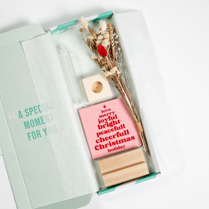 Roze kersttegel met in het rood gelukswensen in kerstboomvorm geschreven met een vaasje droogbloemen ernaast. Beiden staan in een houten blokje