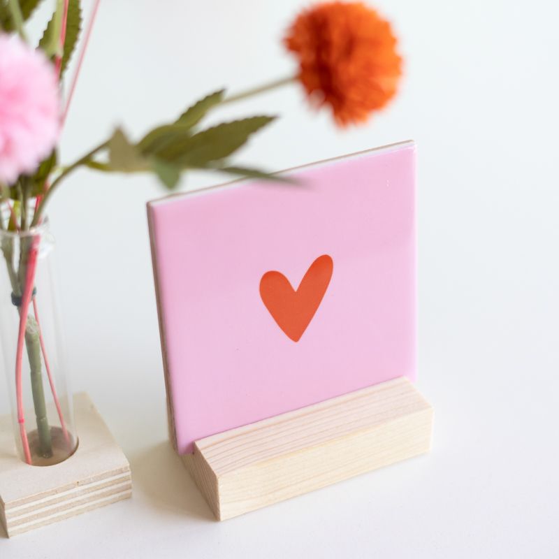 Roze tegeltje met in het midden een klein oranje hartje en het tegeltje staat op een houten houdertje